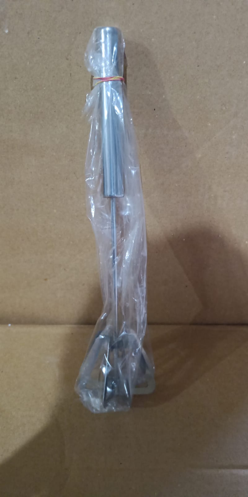 A stainless steel hand whisk wrapped in clear plastic with a red rubber band, against a brown cardboard background.