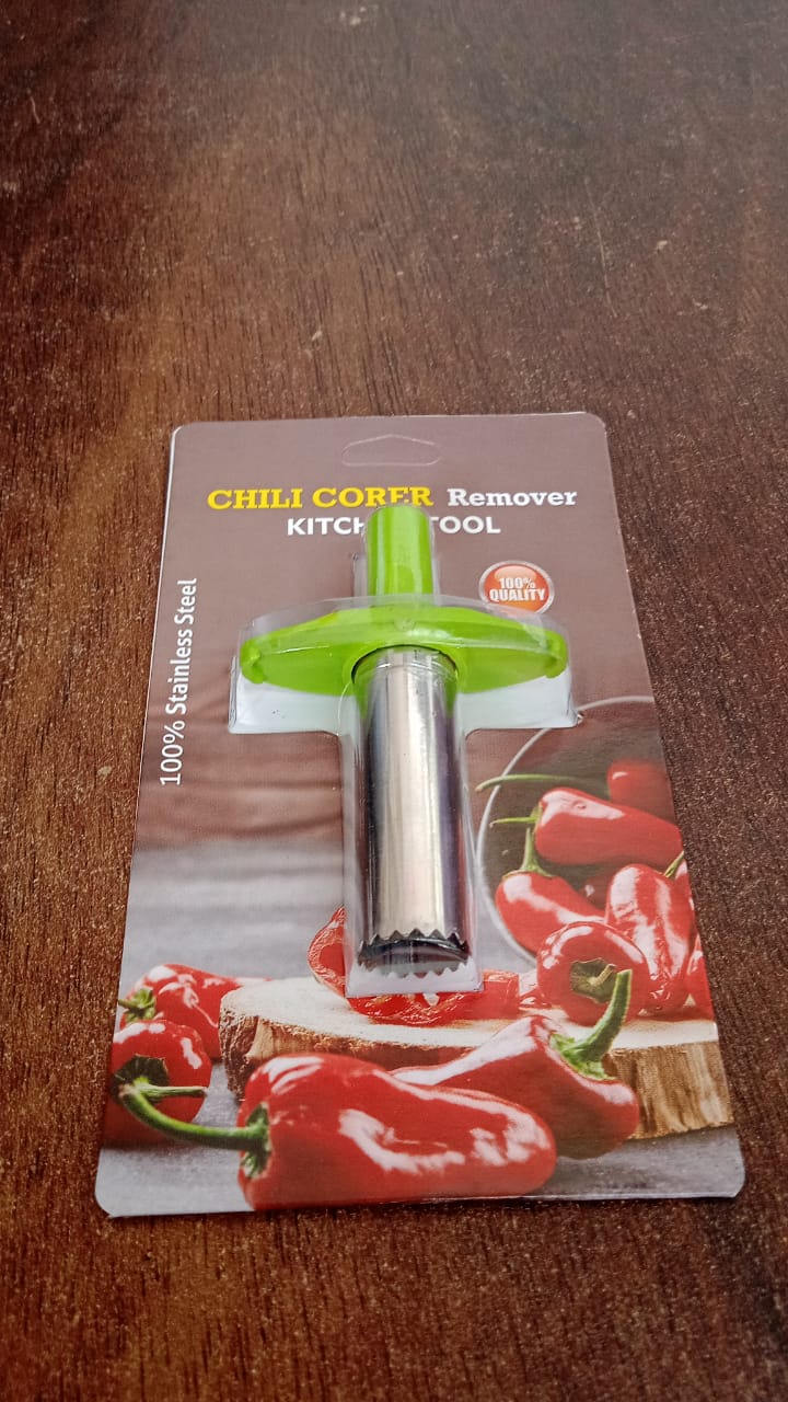 A green and silver chili corer is shown in its retail packaging on a wooden table, featuring red peppers on the card.