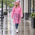 A smiling woman in a pink translucent rain coat with a hood walks on a wet city sidewalk.
