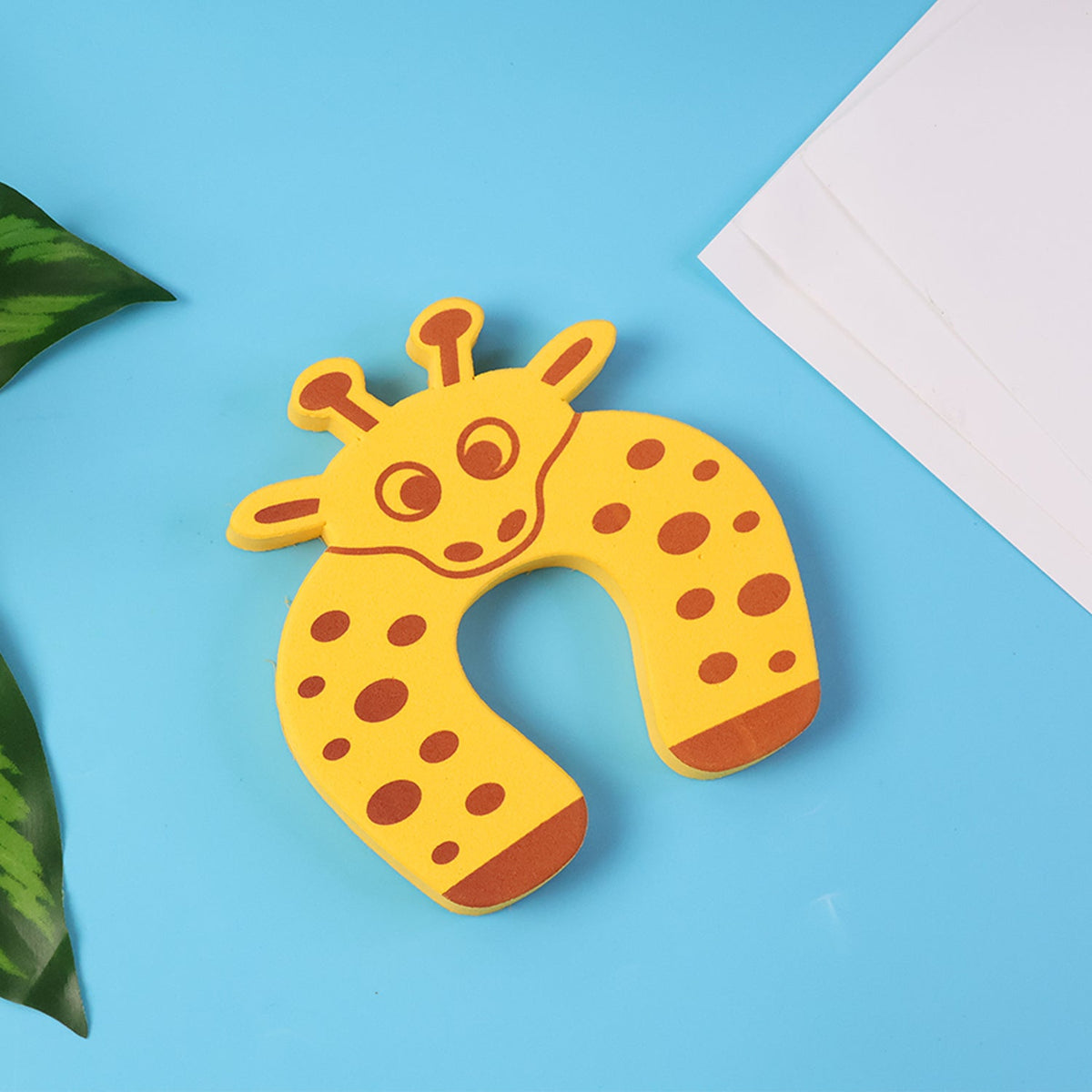 A yellow giraffe-shaped EVA door stopper lies on a blue surface with green leaves and a white paper in the background.