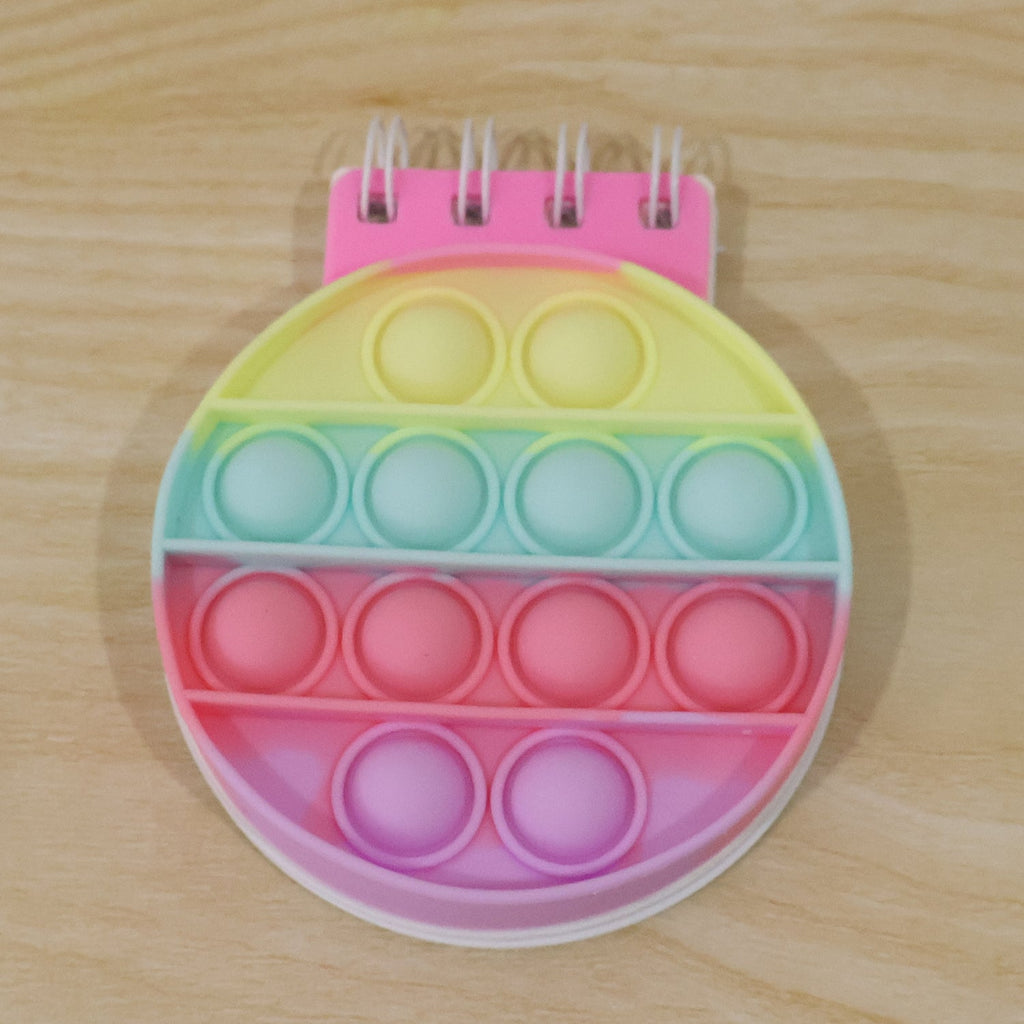 A rainbow pop-it fidget notebook with a pink spiral binding and pastel colored bubbles on a wooden surface.