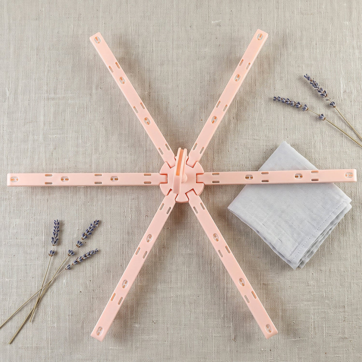 A pink plastic foldable clothes drying rack, unfolded, lies next to a grey cloth and lavender sprigs on a light fabric.