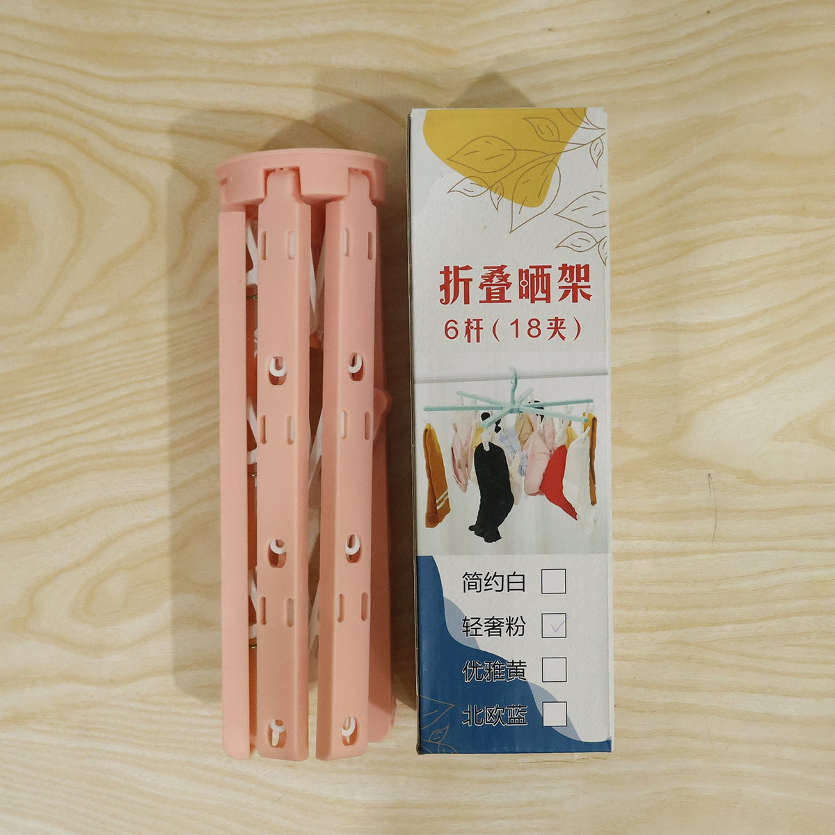A pink plastic foldable clothes drying rack, folded, stands next to its retail box with Chinese text on a wooden surface.