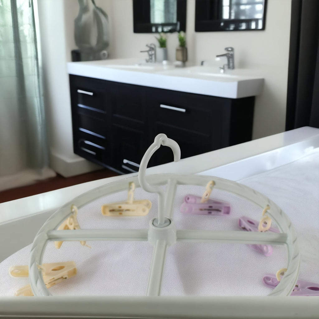 A light blue circular drying rack with purple and yellow clips rests on a white surface in a bathroom setting.