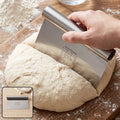A hand uses a stainless steel dough scraper to divide a large round of dough on a wooden cutting board.