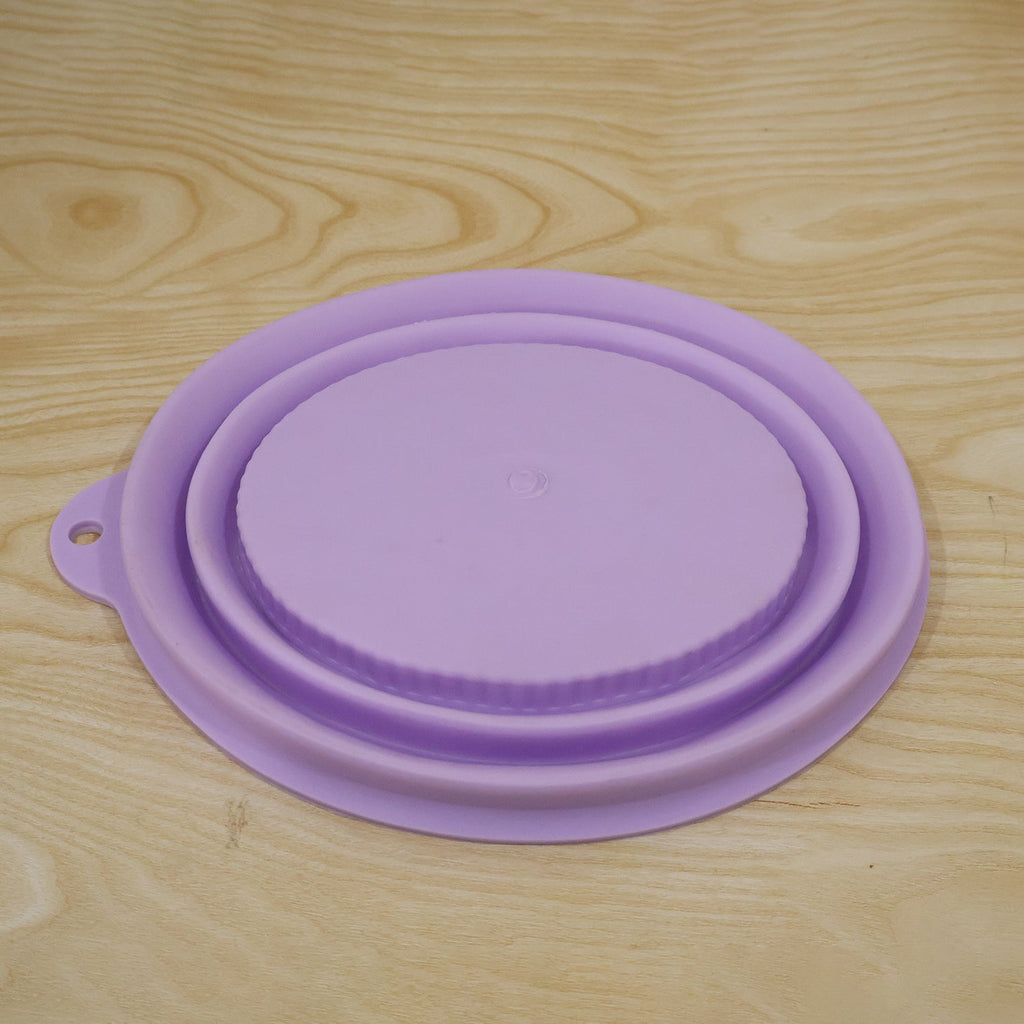 A fully collapsed purple silicone bowl with a textured lid lies flat on a light wood surface.