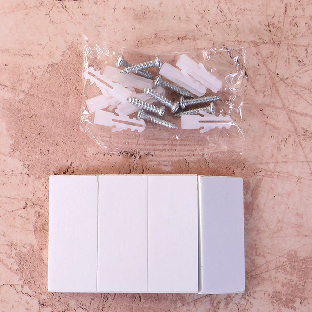 A clear bag of screws and wall anchors sits above a stack of white adhesive strips on a textured surface.