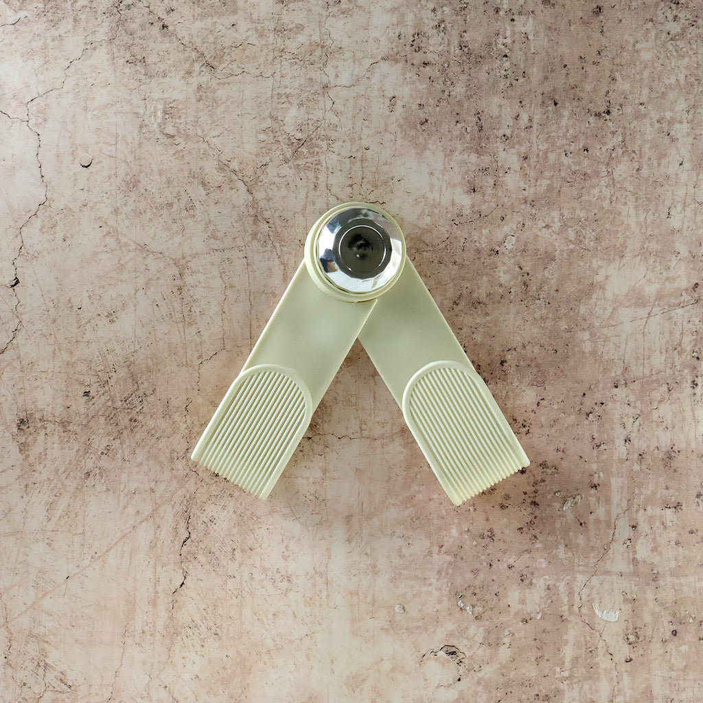 A cream-colored, V-shaped pot lid holder with a chrome center is displayed against a textured, light brown background.