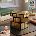 A gold mirror cube puzzle sits on a dark brown desk next to books and a lamp, with a small inset of a child playing with the
