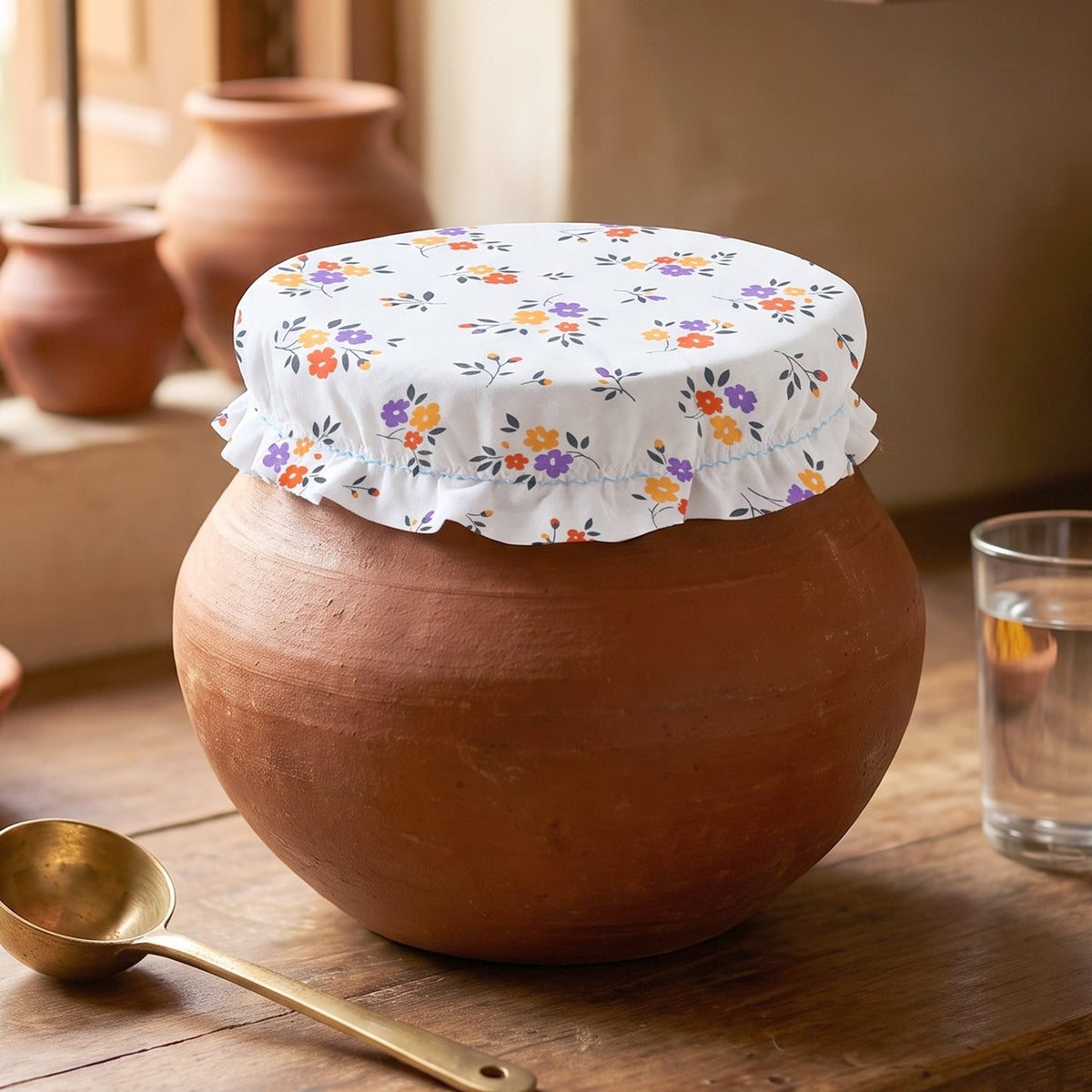Floral food cover on a terracotta pot, with a brass spoon and glass of water nearby.