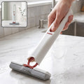 A hand holding a white and red mini mop with a grey sponge head, cleaning a wet white countertop.