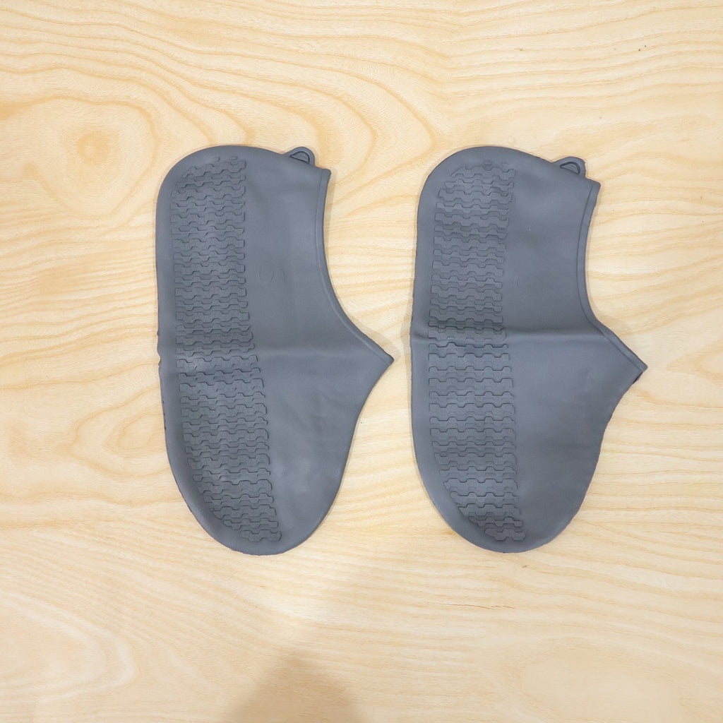Two dark grey silicone shoe covers with textured soles are laid flat on a light wooden surface.
