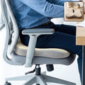 A person sitting on a grey office chair with a beige and grey ergonomic seat cushion