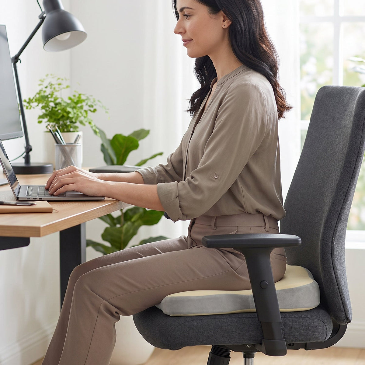 A woman sitting at a desk on a grey office chair with a beige and grey ergonomic seat cushion, typing on a laptop.