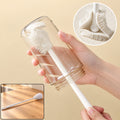 A hand holds a clear glass bottle being cleaned by a white silicone brush, with a close-up of the brush head.