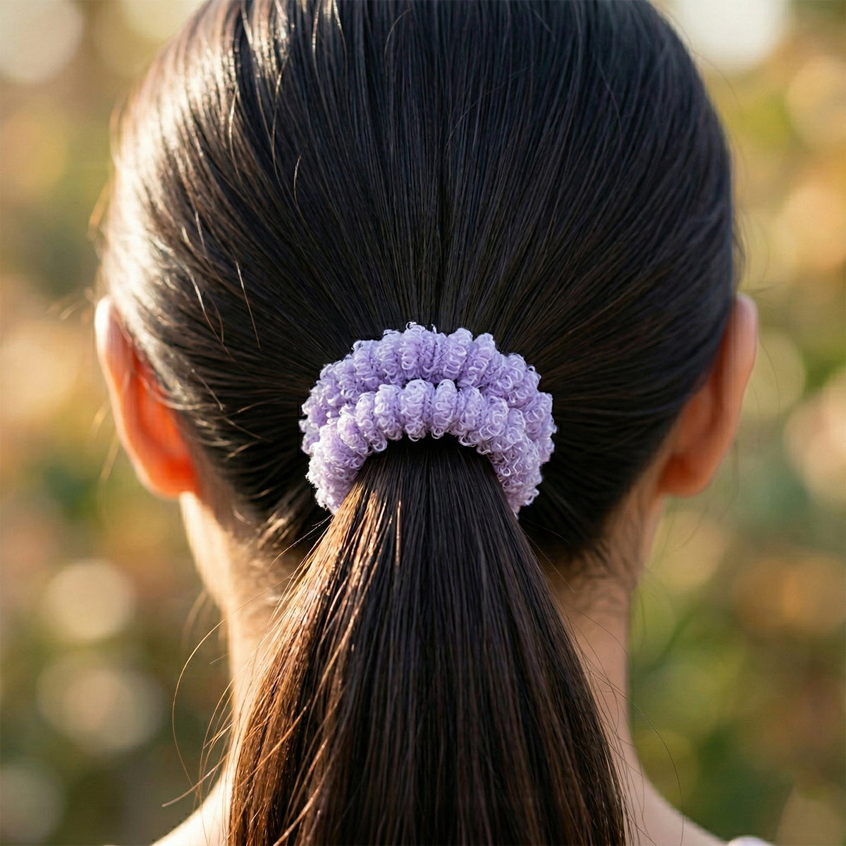 A close-up of a child's dark hair tied in a ponytail with a purple fluffy elastic hair band against a blurred outdoor