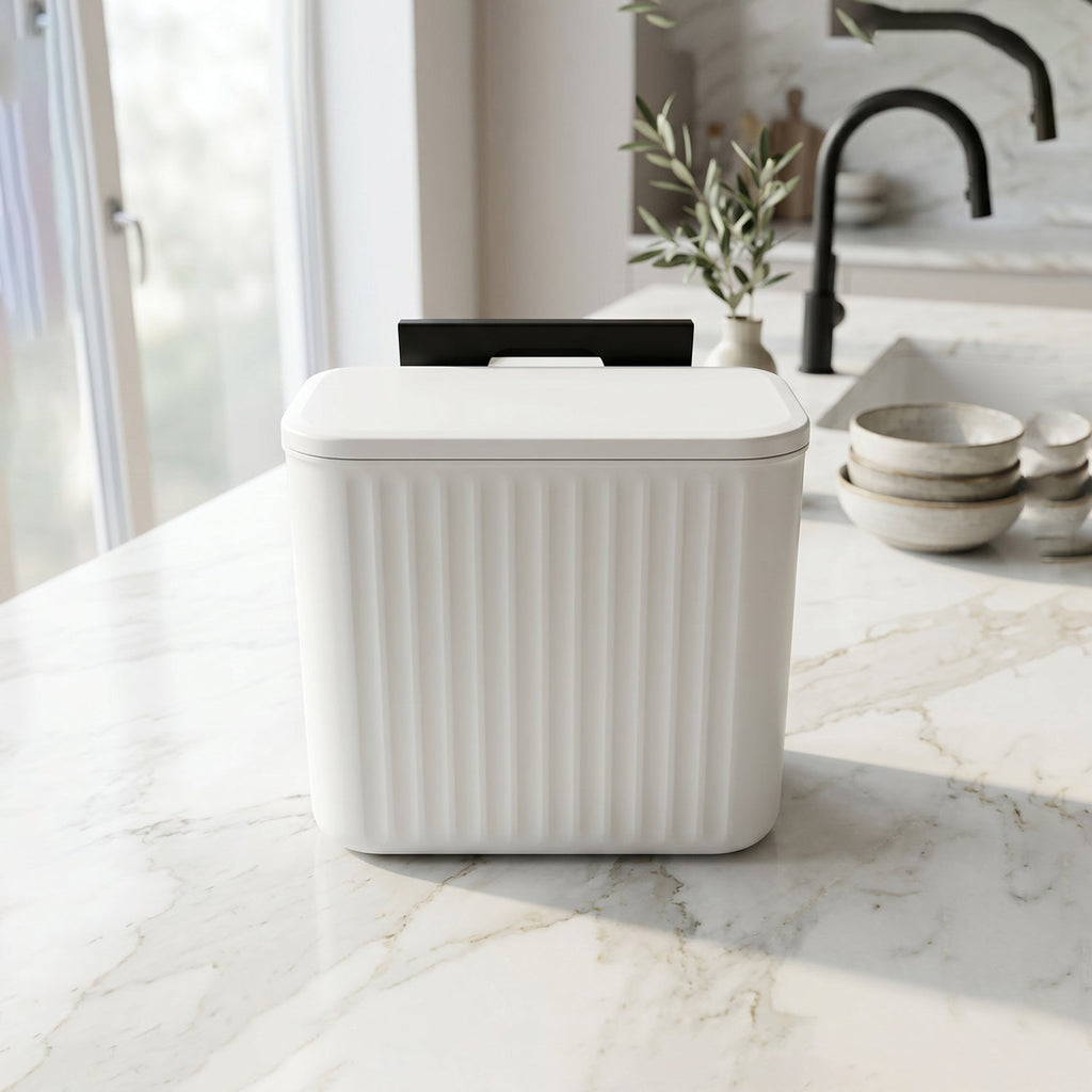 A white ribbed compost bin with a closed lid and black mounting bracket sits on a white marble kitchen counter.