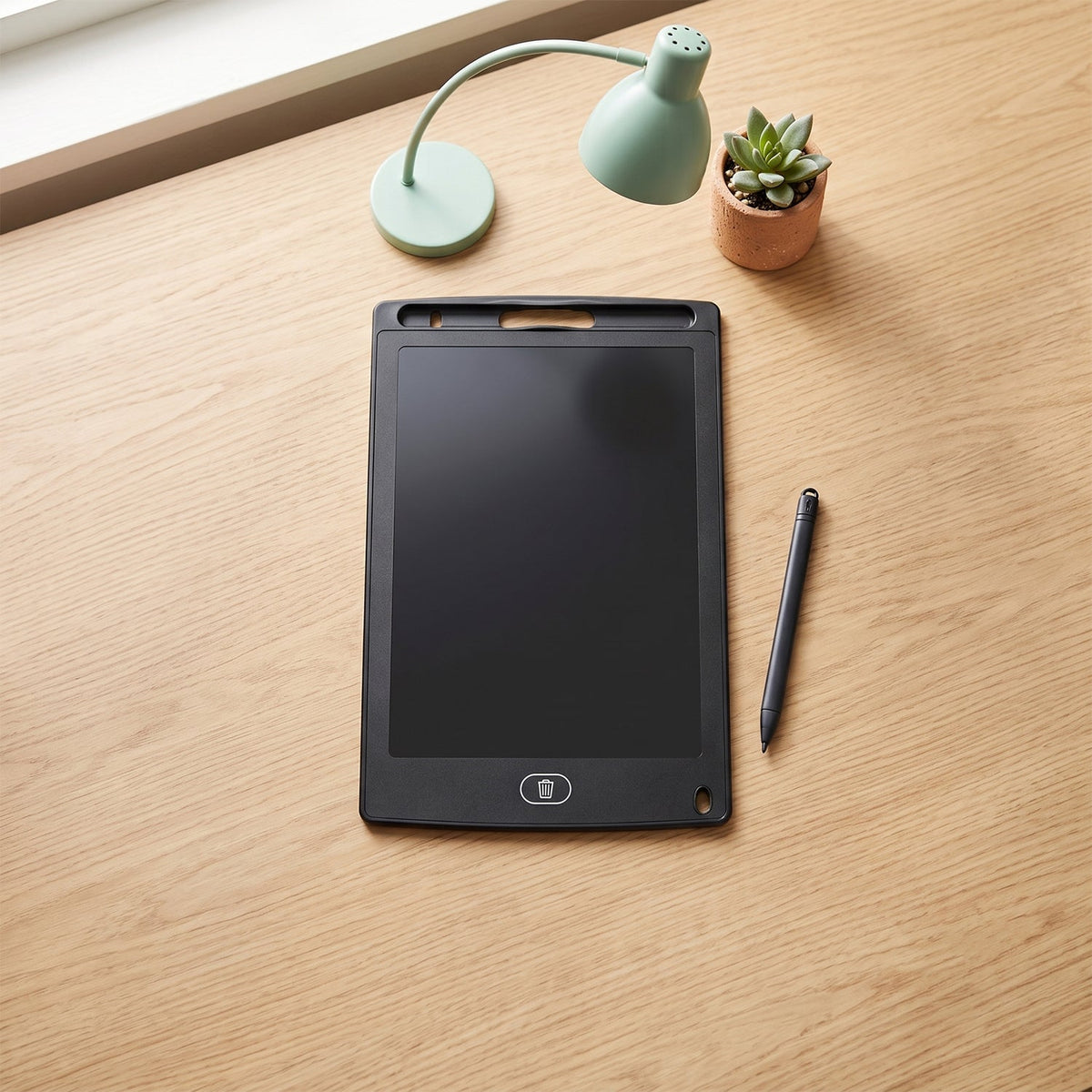 A black LCD writing tablet and stylus rest on a light wooden desk next to a mint green lamp and a small potted plant.