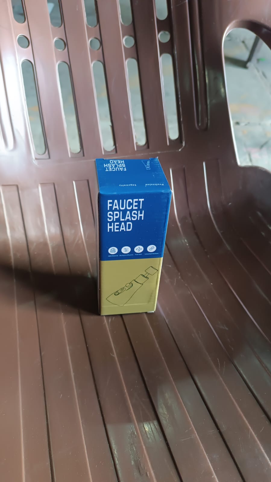A blue and tan box labeled "FAUCET SPLASH HEAD" sits on a brown slatted chair.