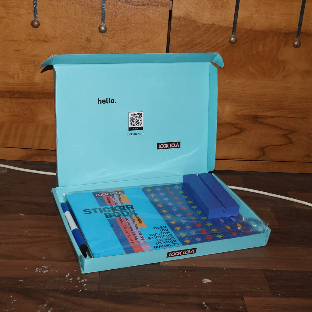 An open light blue box containing a sticker book, blue magnetic strips, and a pen, on a wooden floor.