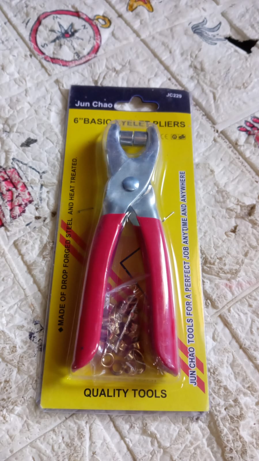 Red-handled grommet pliers in yellow packaging with text "Jun Chao 6" Basic Eyelet Pliers" on a patterned surface.