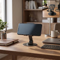 A black metal mobile stand holds a phone horizontally on a wooden desk with a laptop and coffee mug.