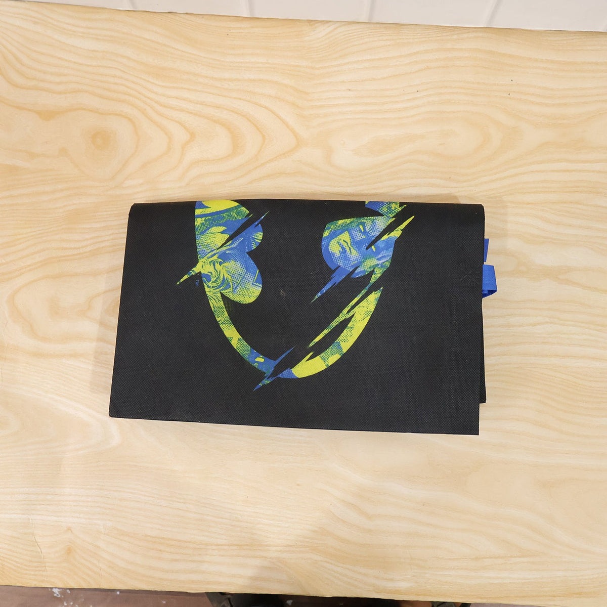 Top-down view of a folded black reusable tote bag with a distressed neon green and blue Batman logo on a light wood surface.