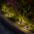 Four silver solar ground lights illuminate a garden bed with purple flowers and green foliage at night.